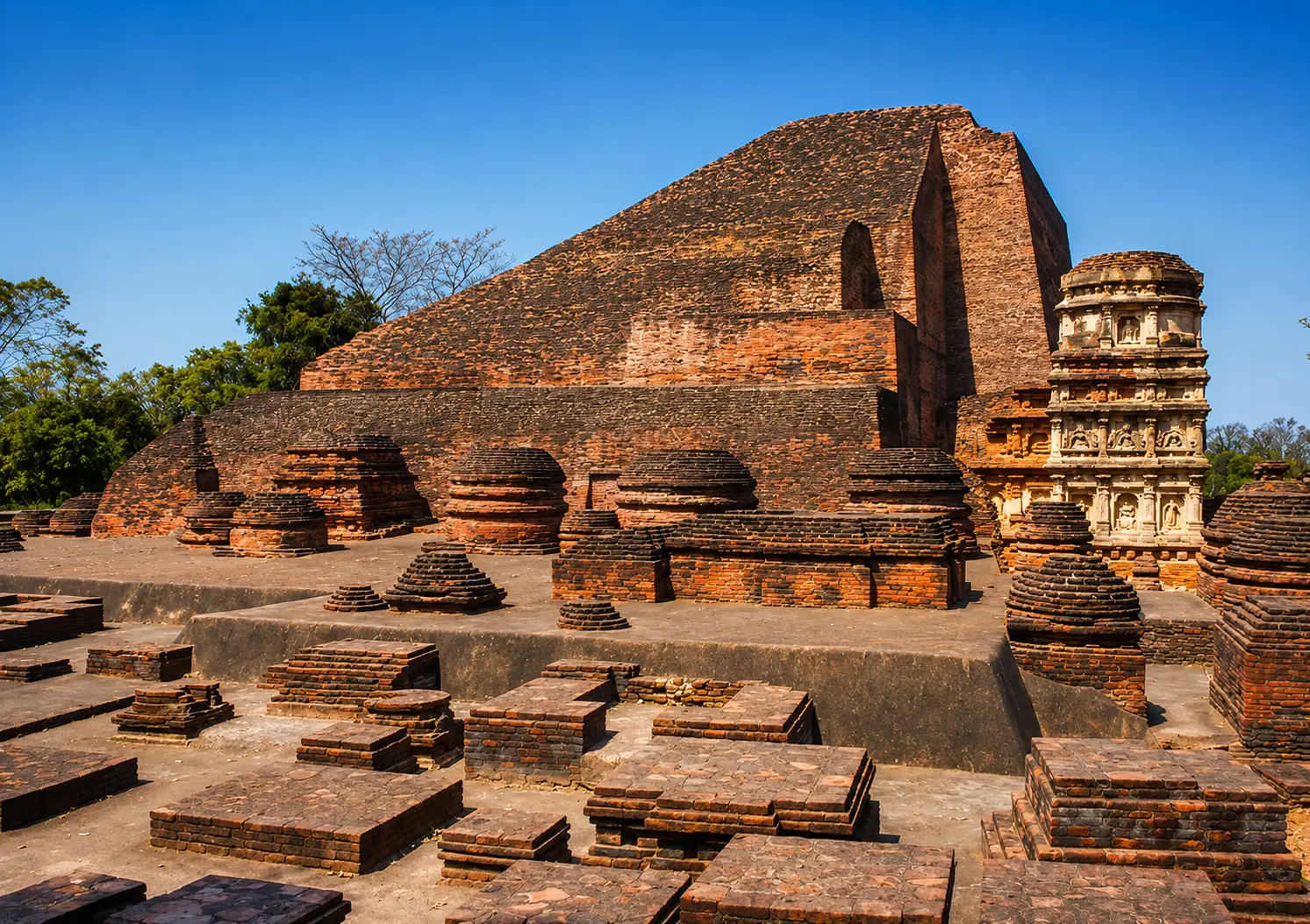नालंदा महाविहार के प्राचीन ईंट-निर्मित खंडहर, जिनमें एक विशाल मंदिर संरचना, छोटे स्तूप और पुरातात्विक अवशेष साफ़ नीले आकाश के नीचे दिखाई दे रहे हैं।